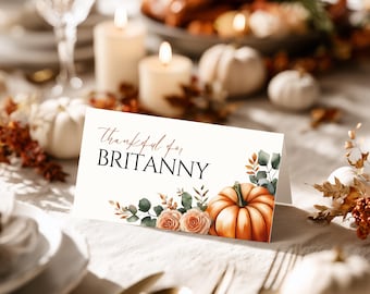 May include: A place card with the text "Thankful for BRITANNY" in black and brown script. The card features a watercolor design of a pumpkin, roses, and greenery, set on a table with candles and pumpkins.