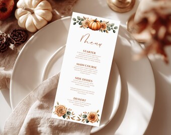 Könnte beinhalten: Eine weiße Menükarte mit Kürbis- und Blumendesign, platziert auf einem weißen Teller mit einer beigen Serviette. Das Menü zeigt das Wort "Menu" in handschriftlicher Schrift und listet Vorspeise, Hauptgericht, Beilagen und Dessert auf.