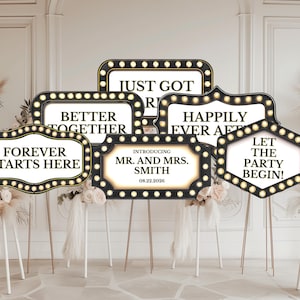 May include: A collection of black and gold wedding photo booth props with phrases like "Forever Starts Here" and "Let the Party Begin!" The props are on gold stands and decorated with lights.