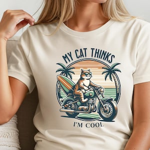 以下が含まれることがあります： サングラスをかけた猫がサーフボードを積んだバイクに乗っている漫画のイラストが描かれた、クリーム色のTシャツ。「MY CAT THINKS I'M COOL」の文字とヤシの木のイラストが円形に配置されています。