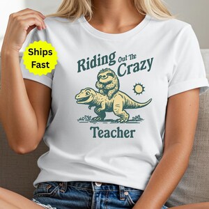 May include: White t-shirt featuring a whimsical illustration of a sloth riding a dinosaur, with the text "Riding Out The Crazy Teacher". The design is in shades of green and yellow, with a small sun graphic.