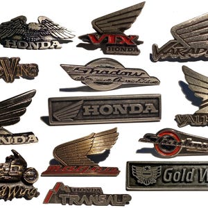 Peut inclure: Une collection de 12 badges de moto en argent et en or avec différents noms de modèles de moto Honda, dont Gold Wing, Vix, Varadero, Valkyrie, Shadow et Transalp.