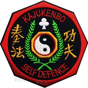 Puede incluir: Parche bordado con fondo negro y borde rojo. El parche presenta el texto "KAJUKENBO SELF DEFENCE" en blanco y amarillo. Un símbolo de yin-yang está en el centro, rodeado por una corona verde y un símbolo de trébol blanco.