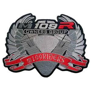 Op de afbeelding: Geborduurde patch met de tekst "M109R OWNERS GROUP" en "m109RIDERS". De patch heeft een zilver en grijs ontwerp met rode accenten, waaronder een banner. De patch heeft de vorm van een schild met vleugels.