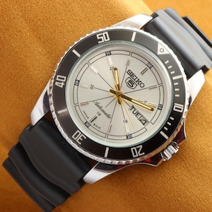 May include: A Seiko 5 automatic watch with a silver face, black bezel, and black rubber strap. The watch face shows the day and date, with gold hands and markers. The watch is marked "Japan Made".