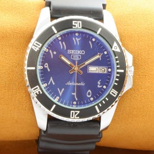 May include: A blue-faced Seiko automatic wristwatch with Arabic numerals and a black bezel. The watch has a date window and a black rubber strap.
