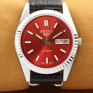 Peut inclure: Une montre-bracelet argentée avec un cadran rouge et un bracelet en cuir noir. Le cadran de la montre a des aiguilles argentées, des index argentés et une fenêtre de date qui affiche "SAT 21". Le cadran de la montre porte également le texte "SEIKO AUTOMATIC JAPAN MADE".