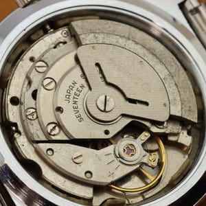 May include: Close-up of a watch mechanism with visible gears, screws, and a balance wheel. The watch's inner workings are exposed, revealing intricate details of its mechanical components. The text "JAPAN SEVENTEEN" is visible on a circular component.