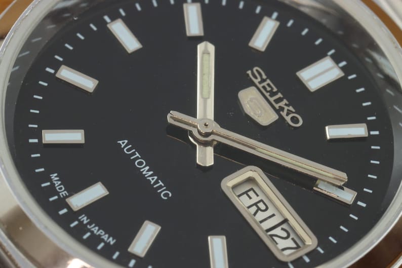 May include: Close-up of a Seiko automatic wristwatch with a black dial. The watch face features silver hands and markers, with the day and date displayed. The text "SEIKO", "AUTOMATIC", "MADE IN JAPAN", and "FRI 27" are visible.