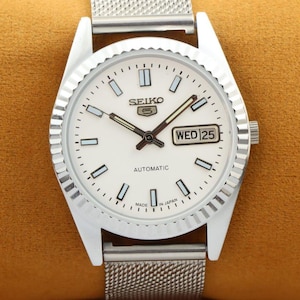 May include: A silver Seiko 5 automatic watch with a white face. The watch has a day and date display, a textured bezel, and a mesh strap. The watch hands and hour markers are silver and black.
