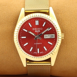 May include: A gold-tone wristwatch with a red face and a gold metal band. The watch face has white markings and the text "SEIKO AUTOMATIC" and "SAT 2".