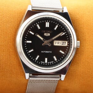 May include: A silver-toned Seiko 5 automatic wristwatch with a black dial. The watch face displays the day and date, with the day reading "FRI 27". The watch has a silver mesh band and is set against a golden-yellow background.
