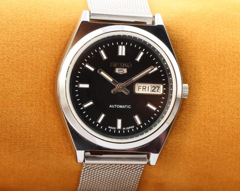 May include: A silver-toned Seiko 5 automatic wristwatch with a black dial. The watch face displays the day and date, with the day reading "FRI 27". The watch has a silver mesh band and is set against a golden-yellow background.
