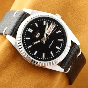 May include: A silver and black automatic wristwatch with a black leather band. The watch face has a day and date display with the words "SUN 13" visible. The watch face also has the word "SEIKO" and "AUTOMATIC" printed on it.
