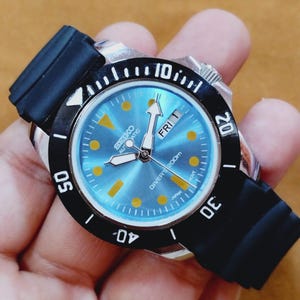Op de afbeelding: Een Seiko automatische duikershorloge met een zwarte lunette en band. De wijzerplaat is lichtblauw met gele markeringen en wijzers. De dag van de week wordt om 3 uur weergegeven. Waterdicht tot 200m.