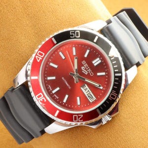 Könnte beinhalten: Eine Seiko 5 Automatik-Uhr mit rotem Zifferblatt und schwarzer und roter Lünette. Die Uhr hat ein silberfarbenes Gehäuse und ein schwarzes Kautschukarmband. Das Zifferblatt zeigt den Tag und das Datum an.