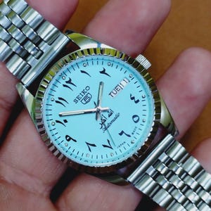 Puede incluir: Un reloj automático Seiko 5 plateado con esfera azul claro. El reloj presenta números arábigos, agujas plateadas y una pantalla de día/fecha. La correa del reloj es de eslabones plateados.