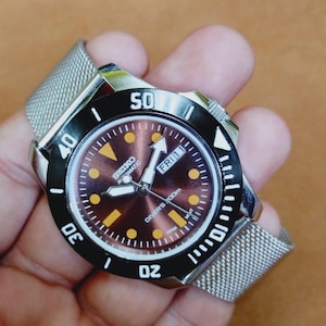 May include: A stainless steel Seiko automatic diver's watch with a brown dial, black bezel, and a mesh band. The watch face features yellow hour markers and the day is displayed as "FRI 11".