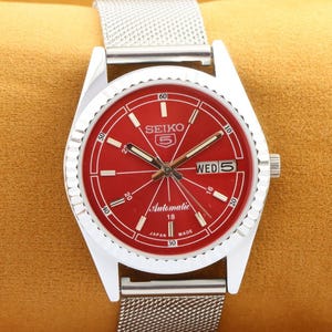 Puede incluir: Un reloj de pulsera plateado con una esfera roja y una correa de malla plateada. La esfera tiene marcas blancas y el texto "SEIKO 5 Automatic 18 JAPAN MADE" y "WED 5".