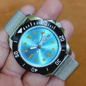 Può includere: Un orologio subacqueo automatico Seiko in acciaio inossidabile con quadrante azzurro. L'orologio presenta una lunetta nera con segni bianchi, un cinturino a maglia e il giorno della settimana visualizzato. Il quadrante ha indici dorati.