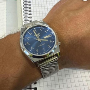 Könnte beinhalten: Eine silberfarbene Seiko Automatik-Armbanduhr mit blauem Zifferblatt und arabischen Ziffern. Die Uhr verfügt über eine Datumsanzeige und ein Metallarmband. Die Uhr ist am Handgelenk.