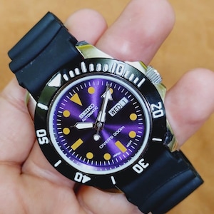 May include: A Seiko automatic diver's watch with a black bezel and a purple dial. The watch has a black rubber strap and features the day of the week. The watch is water resistant to 200m.