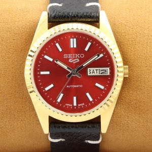 Peut inclure: Une montre-bracelet dorée avec un cadran rouge et un bracelet en cuir noir. Le cadran de la montre comporte des marques blanches et le texte "SEIKO AUTOMATIC JAPAN MADE". Le jour de la semaine est affiché comme "SAT 2".