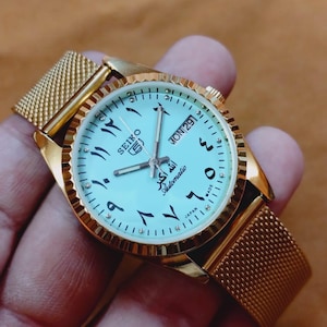 May include: Gold-toned Seiko automatic wristwatch with a light blue face. The watch features Arabic numerals, a day-date display, and a mesh band. The watch face has the text "SEIKO" and "Automatic".