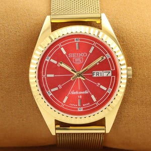 Puede incluir: Un reloj de pulsera automático Seiko 5 dorado con esfera roja. El reloj presenta una correa de malla dorada, agujas doradas y una pantalla de día y fecha. La esfera del reloj incluye el texto "SEIKO 5 Automatic 18 Made Japan".