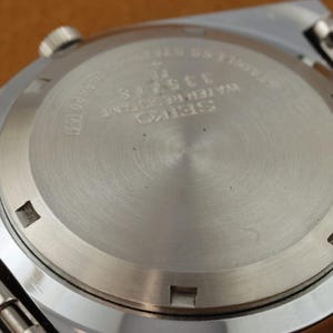 May include: Close-up of the back of a silver-toned Seiko watch. The watch back is circular with etched text including "SEIKO WATER RESISTANT" and the number "335718". The watch band is visible on the sides.