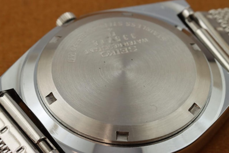 May include: Close-up of the back of a silver-toned Seiko watch. The watch back is circular with etched text including "SEIKO WATER RESISTANT" and the number "335718". The watch band is visible on the sides.