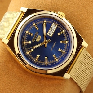 May include: Gold-toned Seiko 5 automatic watch with a blue dial and gold hands. The watch face displays the day and date. The watch has a gold mesh strap.