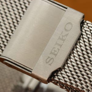 May include: Close-up of a stainless steel watch band with a mesh design. The band features a clasp with the engraved word "SEIKO". The watch band is silver in color and has a woven pattern.