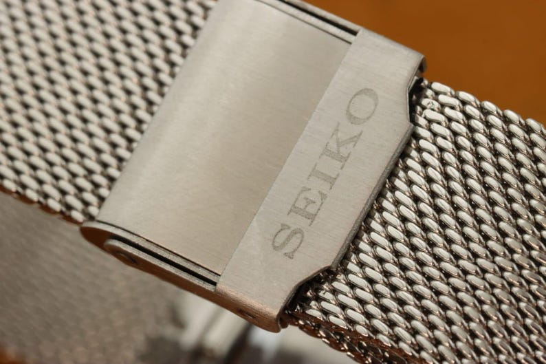 May include: Close-up of a stainless steel watch band with a mesh design. The band features a clasp with the engraved word "SEIKO". The watch band is silver in color and has a woven pattern.