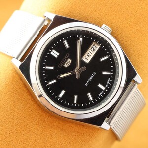 May include: A silver-toned Seiko 5 automatic watch with a black dial. The watch features a day and date display, luminous hands, and a mesh band. The watch face has white hour markers and the text "SEIKO 5 AUTOMATIC MADE IN JAPAN".