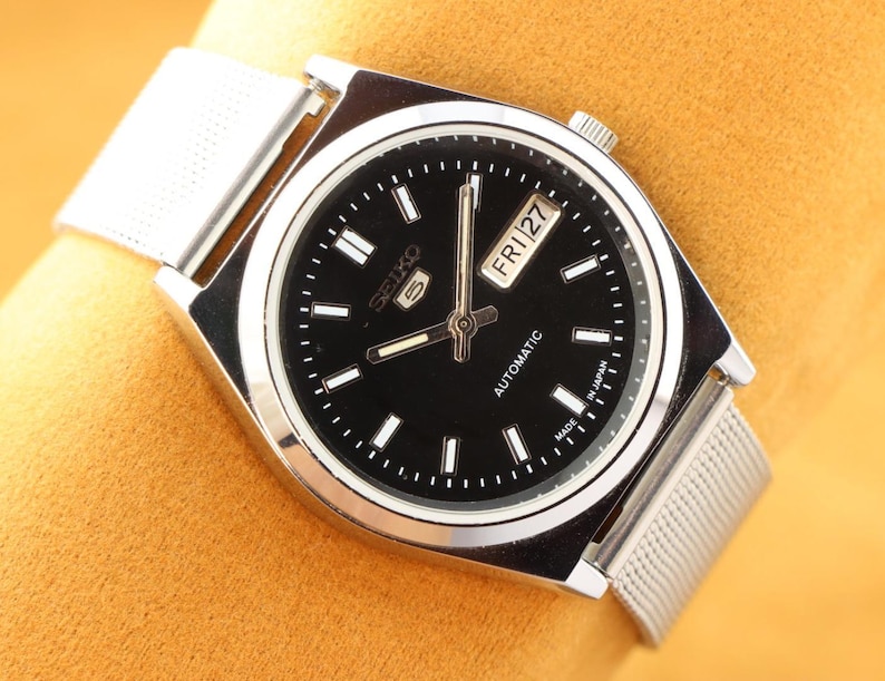 May include: A silver-toned Seiko 5 automatic watch with a black dial. The watch features a day and date display, luminous hands, and a mesh band. The watch face has white hour markers and the text "SEIKO 5 AUTOMATIC MADE IN JAPAN".