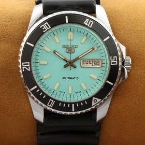 Op de afbeelding: Een zilveren en zwarte Seiko 5 automatische polshorloge met een lichtblauwe wijzerplaat en zwarte wijzers. Het horloge heeft een zwarte bezel met witte markeringen en een zwarte rubberen band.