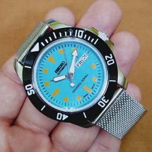 Może przedstawiać: Srebrny zegarek Seiko Automatic Diver's z turkusową tarczą i czarnym pierścieniem. Zegarek posiada bransoletę z siatki, pomarańczowe znaczniki godzin i datę piątek 26. Wodoszczelność do 200m.