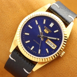 May include: A gold-toned wristwatch with a navy blue face, featuring white hour markers and hands. The watch displays "SEIKO 5 AUTOMATIC" and "WED 31". It has a black leather band with white stitching. The watch is Made in Japan.