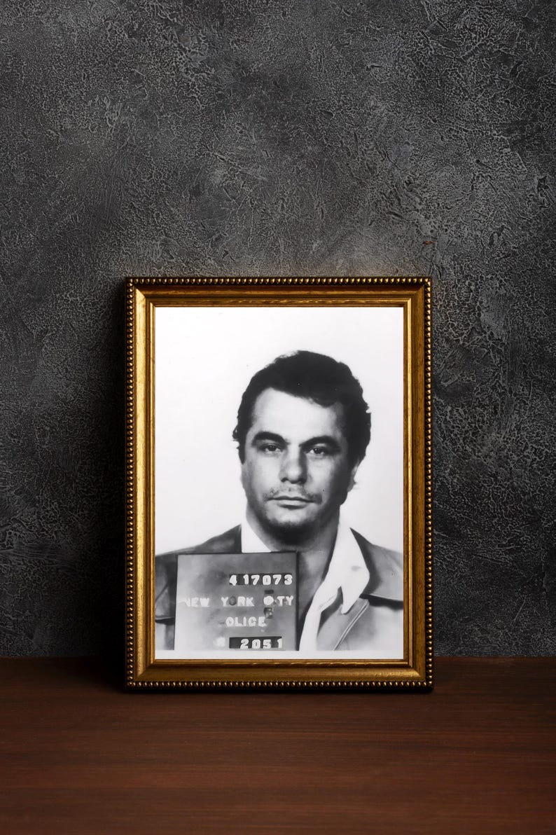 May include: Black and white mugshot of a man in a framed photo. The photo shows the man's face and shoulders. The photo is in a gold frame with a white mat. The photo has the text "4 17073 NEW YORK CITY POLICE 2051" printed on the bottom.