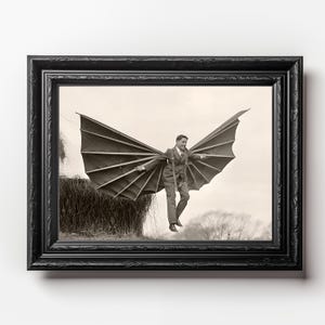 May include: A black and white photograph of a person wearing a suit and a pair of large, homemade wings, flying over a field of hay.