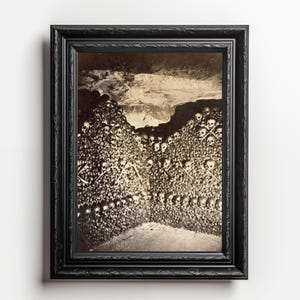 May include: A black and white photograph of a wall made entirely of human skulls and bones. The skulls are arranged in a symmetrical pattern, with the bones forming a cross shape in the center. The photograph is framed in a black frame.