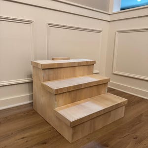 May include: A three-tiered wooden display stand with a natural finish. The stand has a rectangular shape and is designed to showcase items. The steps are approximately 10 inches deep. The stand is placed on a wooden floor.