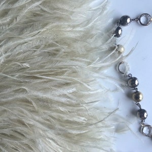 Puede incluir: Un boa de plumas de avestruz blanco con un collar de cuentas plateadas y doradas sobre un fondo blanco.