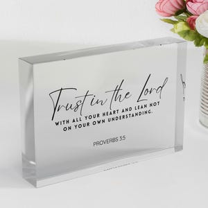 May include: A clear acrylic block with the script "Trust in the Lord" and the Bible verse Proverbs 3:5 in black lettering. The block is rectangular and sits on a white surface, with a bouquet of pink and white flowers in a silver container in the background.