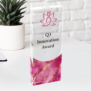 May include: A clear acrylic award with a pink marble design at the base. The award features a pink lotus flower logo and the text "Q3 Innovation Award" in black. A small potted succulent is in the background.