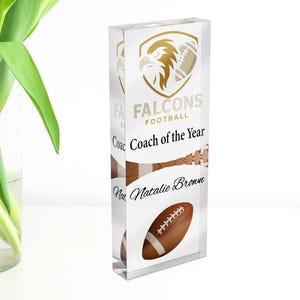 Könnte beinhalten: Eine transparente Acryl-Auszeichnung mit einem goldenen Falcons-Logo und dem Text "Falcons Football Coach of the Year Natalie Brown". Die Auszeichnung zeigt eine Football-Grafik und hat eine rechteckige Form.