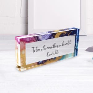 May include: A rectangular acrylic block with a colorful abstract design in shades of pink, purple, gold, and blue. The block features the quote "To live is the rarest thing in the world" by Oscar Wilde.