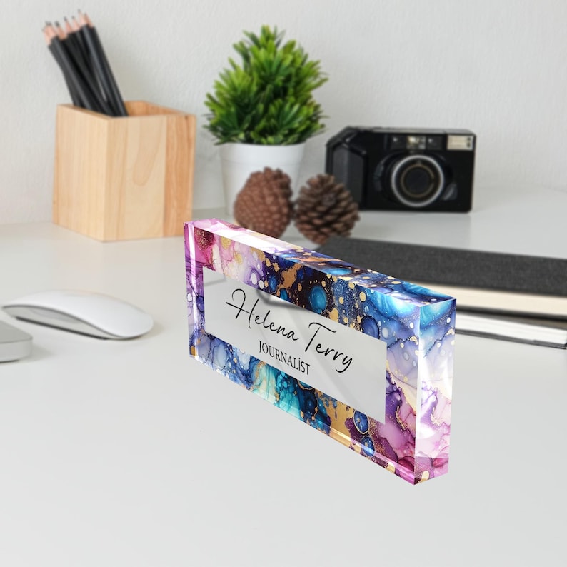 May include: A clear acrylic desk nameplate with a colorful abstract design in shades of blue, purple, and gold. The name "Helena Terry" and the title "Journalist" are printed in black on a white background.