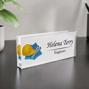 Puede incluir: Una placa de escritorio rectangular de acrílico transparente con el nombre impreso Helena Terry y el título Engineer. A la izquierda del texto hay una ilustración gráfica de un casco de seguridad amarillo sobre un plano arquitectónico azul.